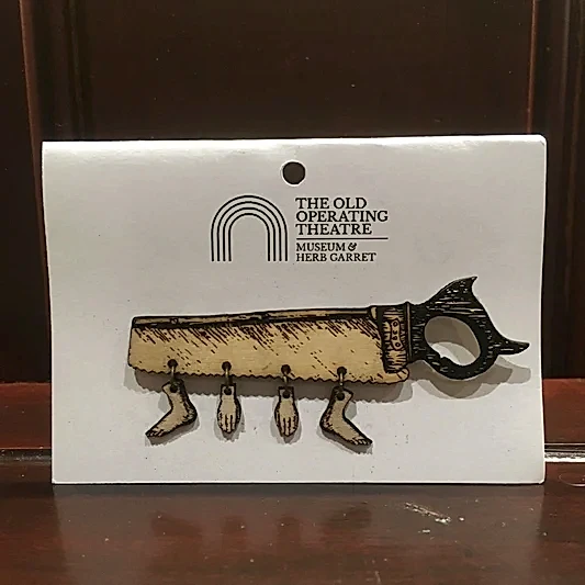 A wooden brooch in the shape of a saw, with two hands and two feet dangling from it